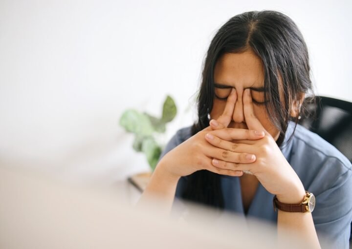 6 tipos de estrés: una mujer está en su oficina y se muestra muy cansada. Toma su rostro con las manos y presiona su frente para aliviar el dolor de cabeza.