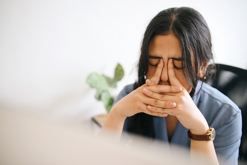 6 tipos de estrés: una mujer está en su oficina y se muestra muy cansada. Toma su rostro con las manos y presiona su frente para aliviar el dolor de cabeza.
